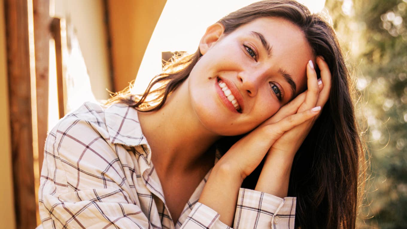A woman smiling and resting her head on her hands outdoors in warm sunlight.