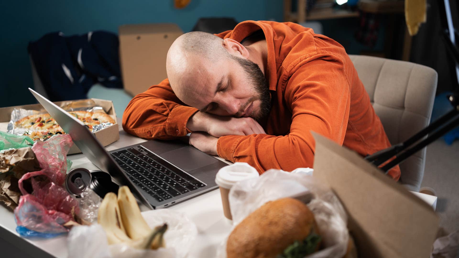 man asleep at computer surrounded by clutter and trash