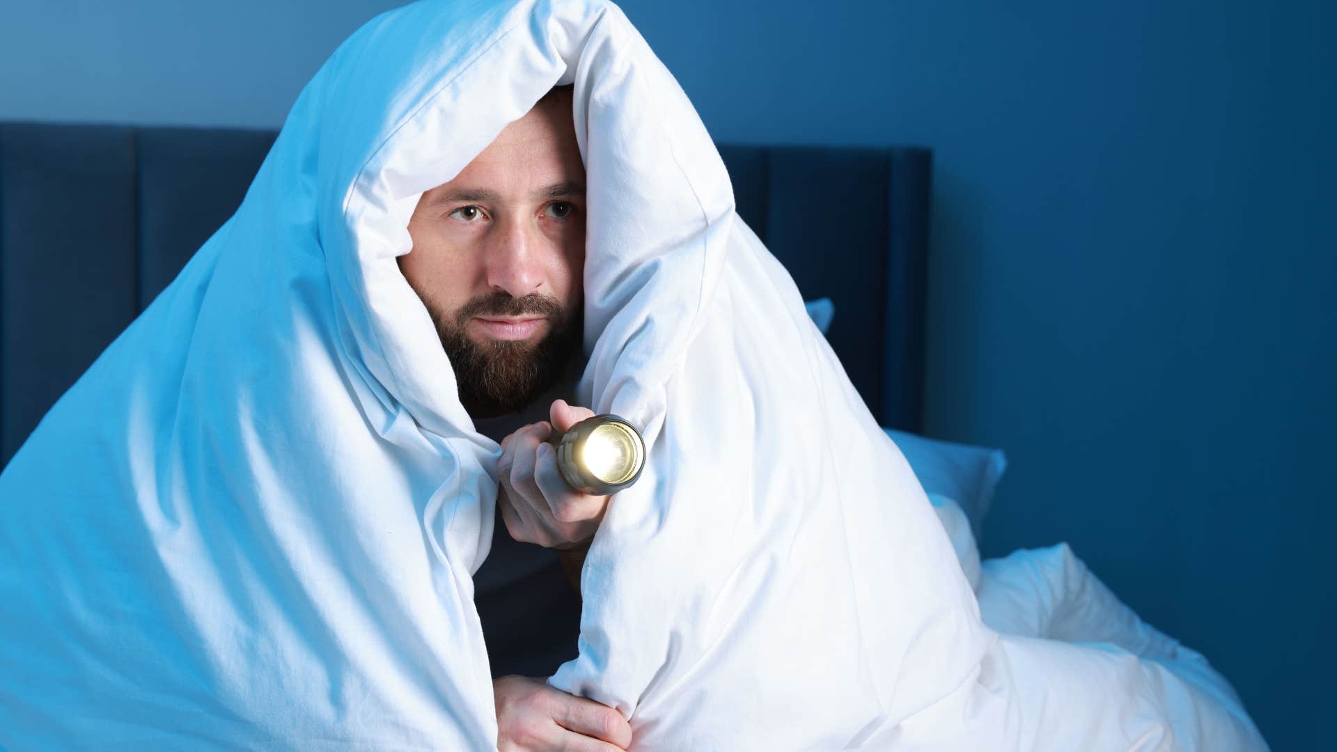 nervous man who's afraid of the dark holding a flashlight in bed
