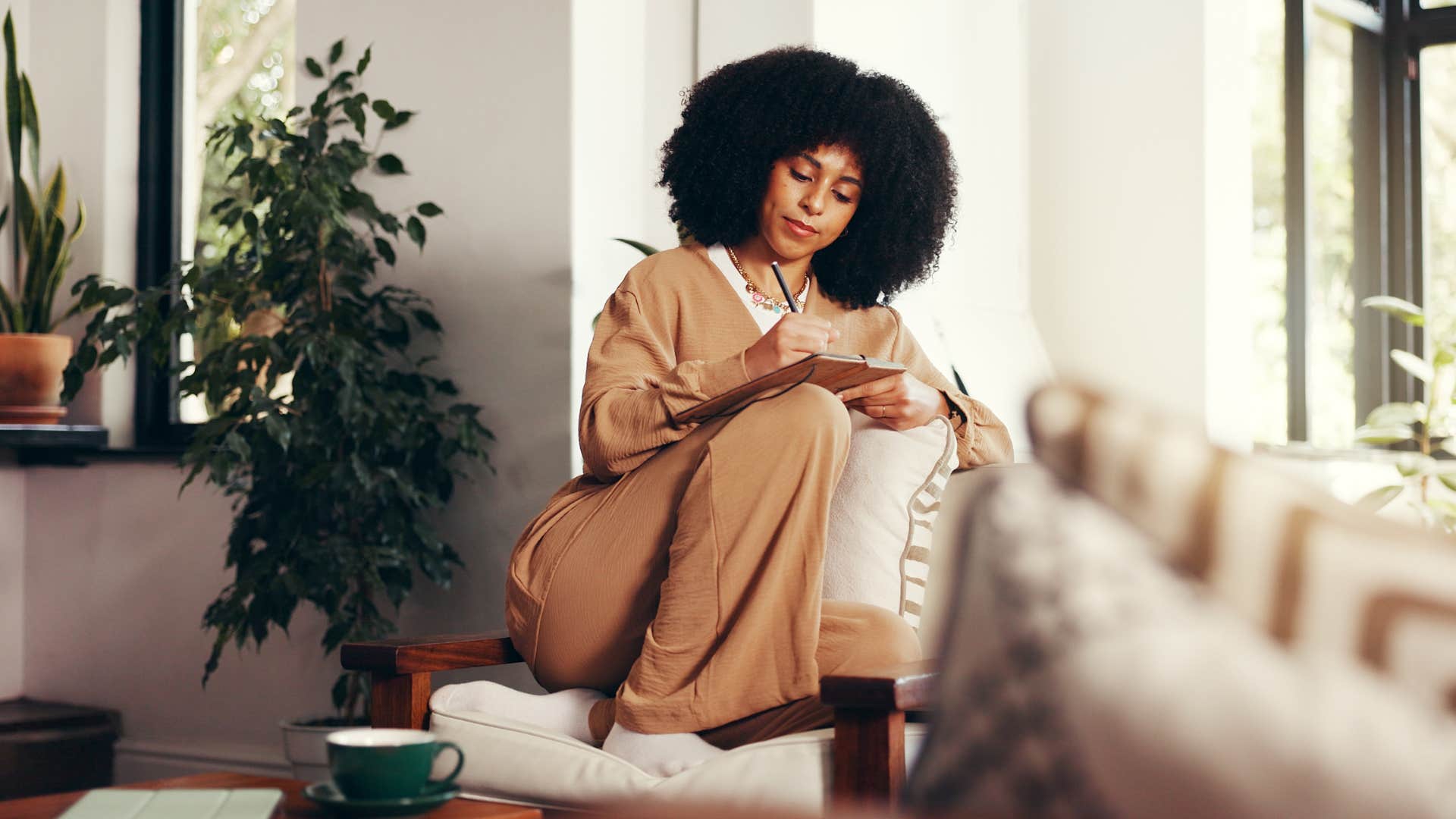 woman writing in notebook sitting on couch by window