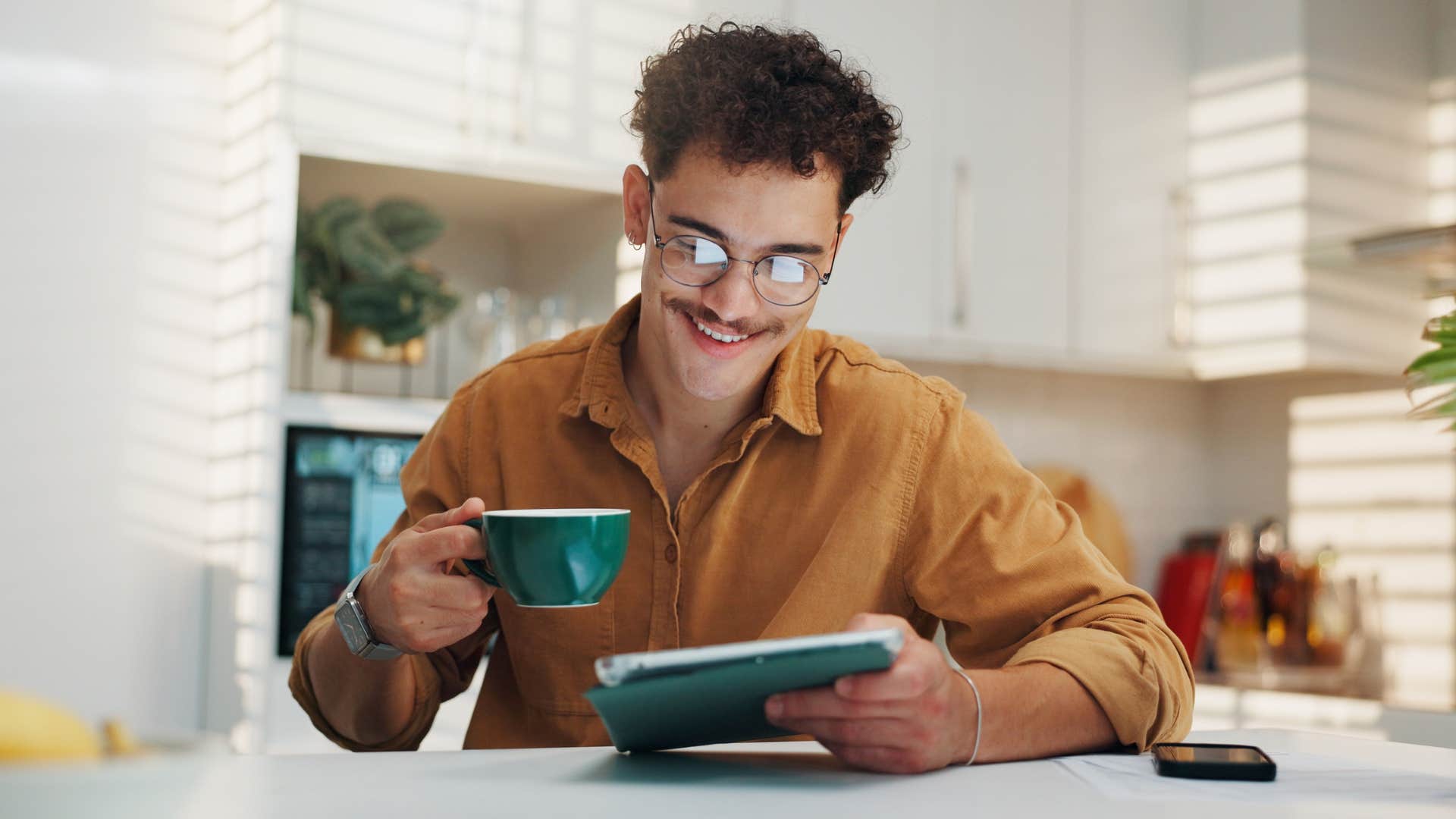 man drinking from mug while reading on tablet