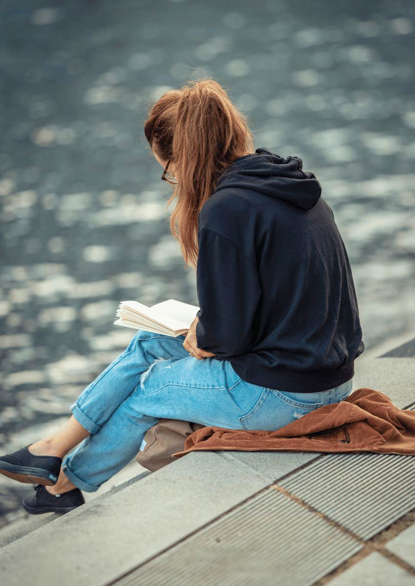 young woman reading book outdoors near lake