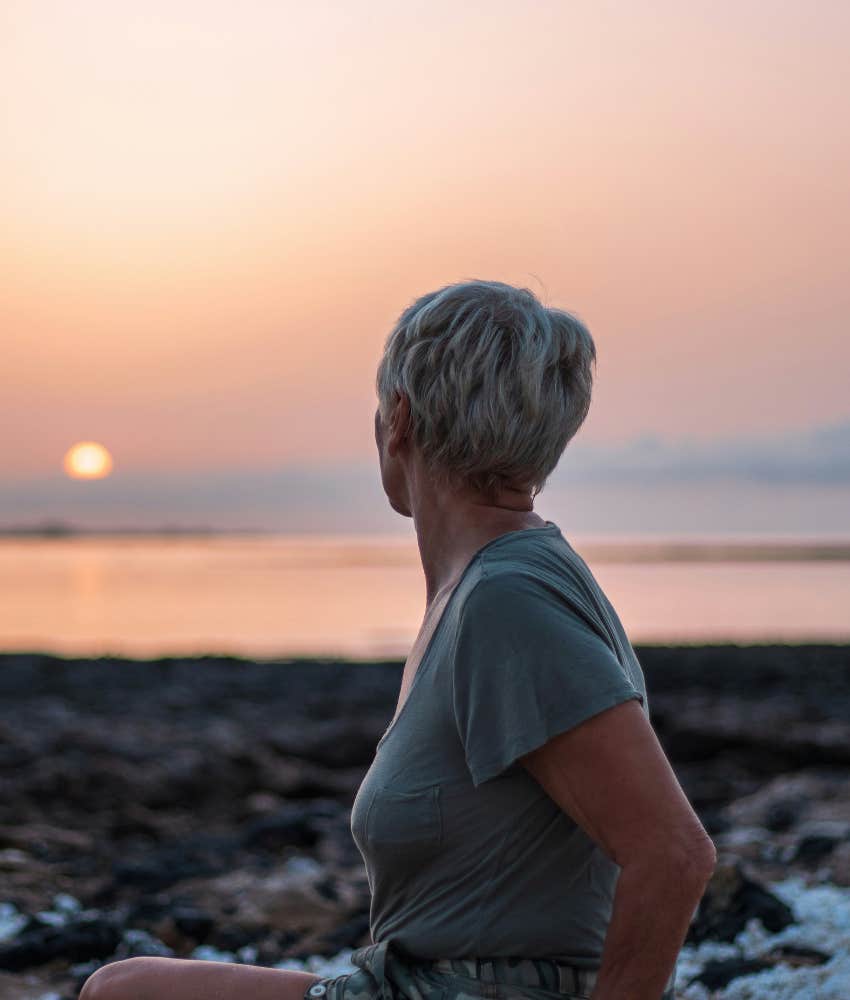 older woman watches sunset into ocean showing impact of cancer diagnosis