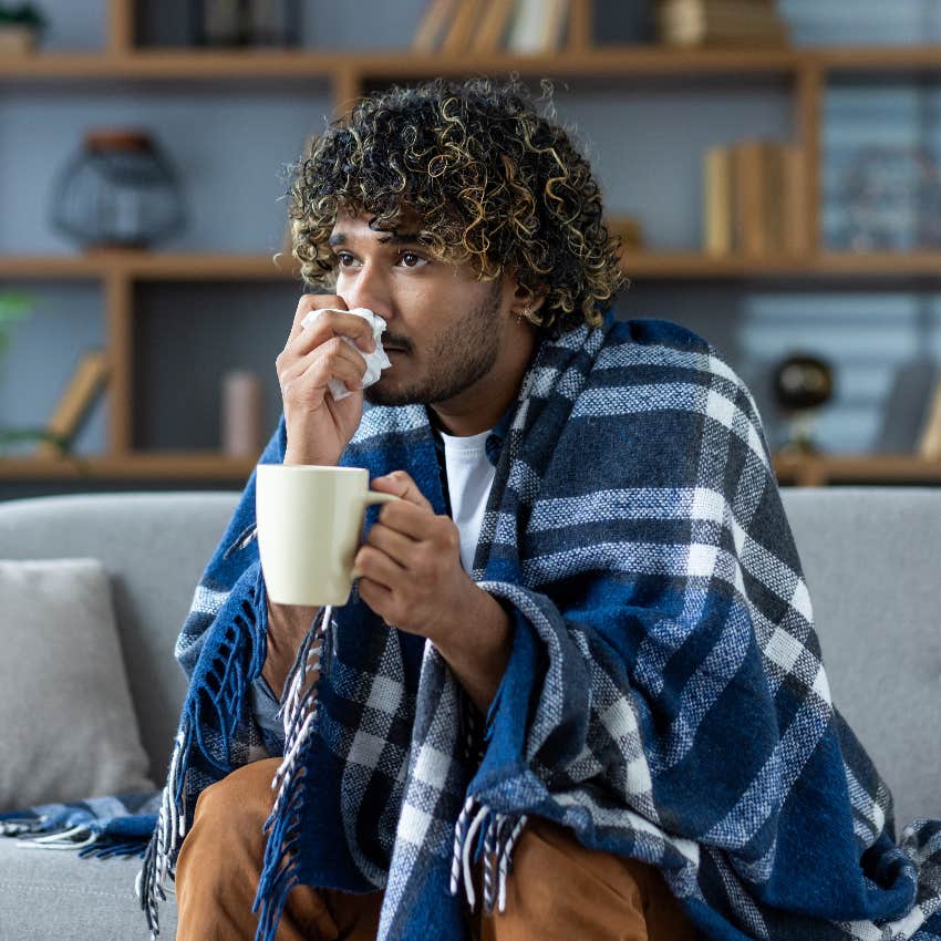 sick man sitting on couch with blanket over shoulders holding mug