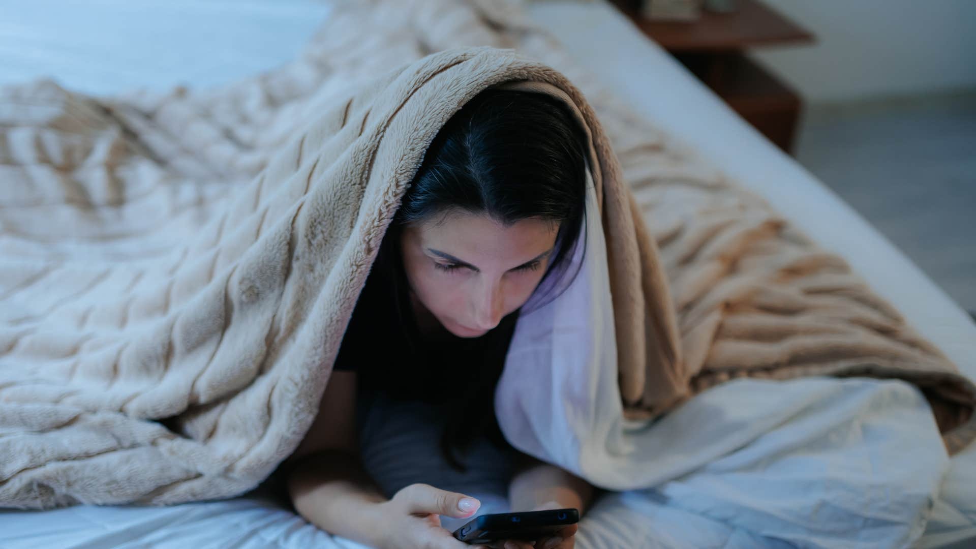 woman under blanket on phone scrolling through social media as she realizes Nobody's life looks as stable as it does online