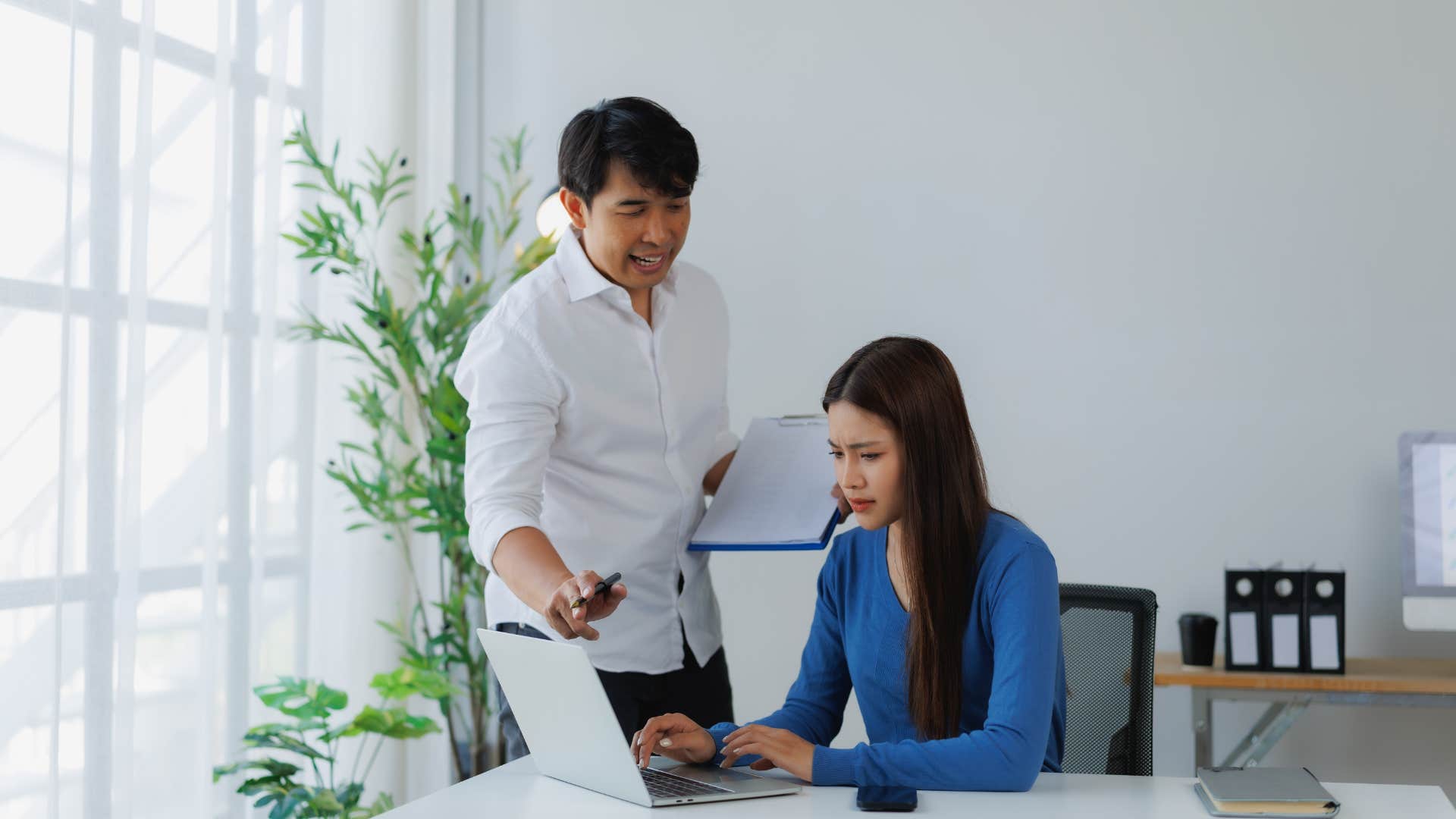 boss hovering over employee working at desk micromanaging her