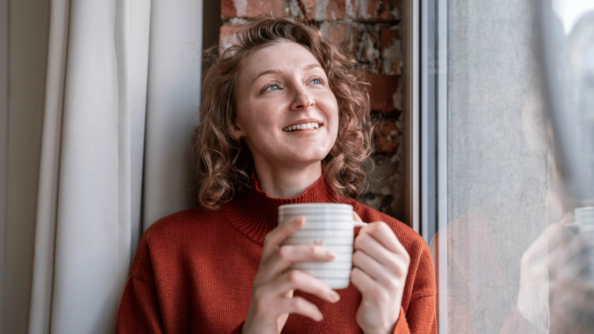 smiling woman standing by window holding mug