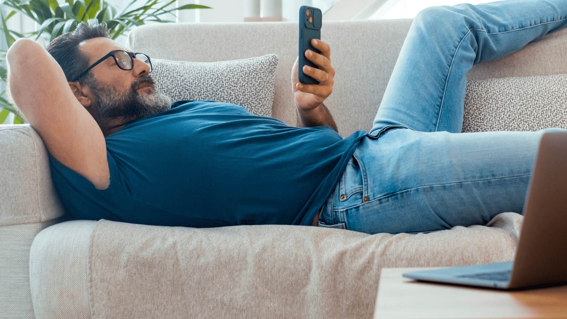 man lazing on couch on phone as he thinks i'll get to it eventually