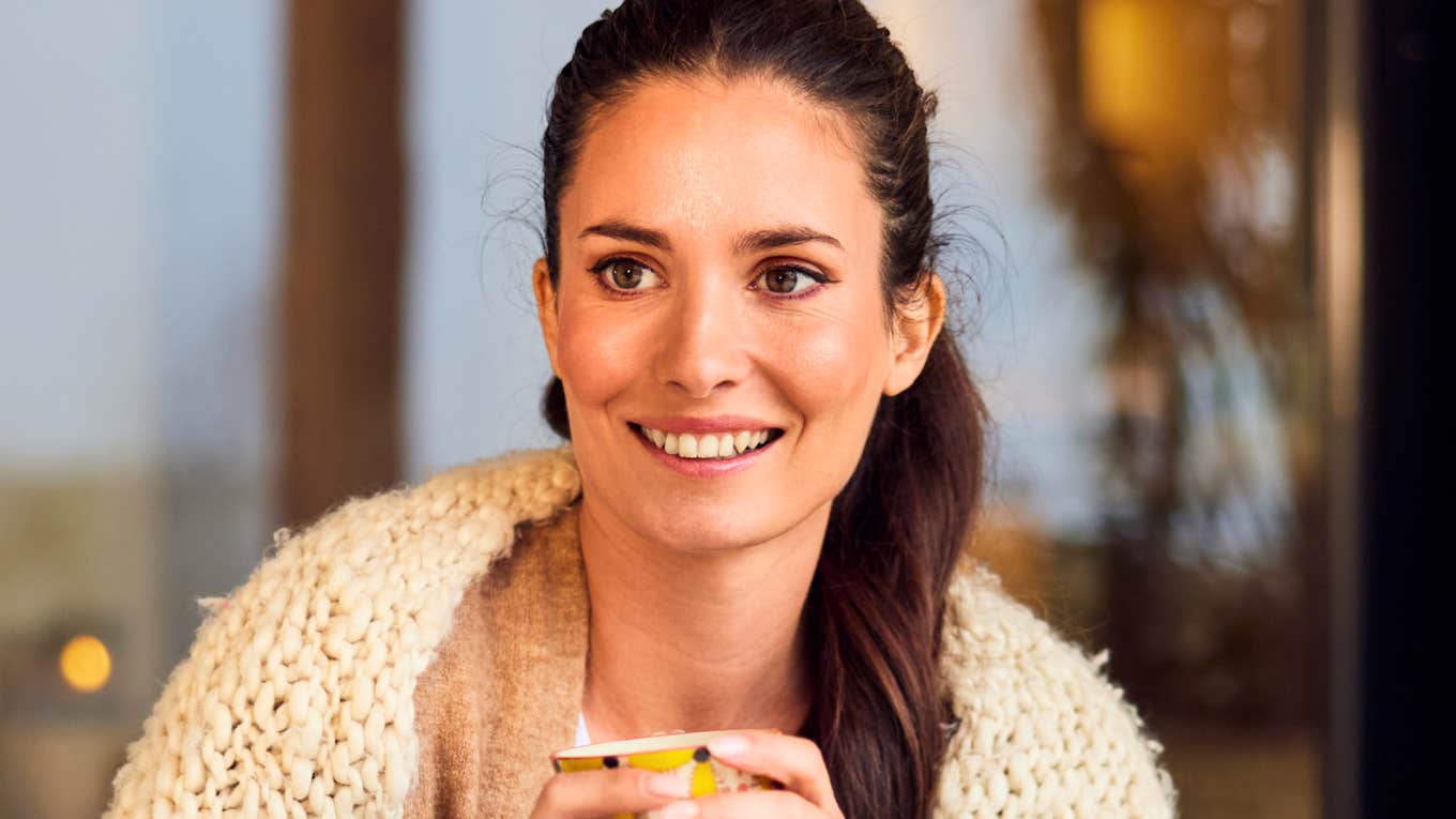 A smiling brunette woman sitting outdoors with her friend on a crisp autumn day, holding a warm cup of tea while enjoying a captivating conversation.