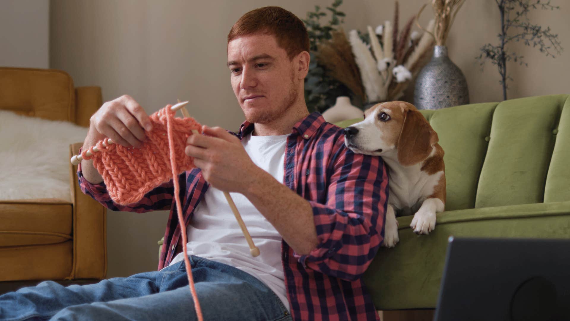 man in white shirt and plaid jacket knitting and crocheting with dog behind him