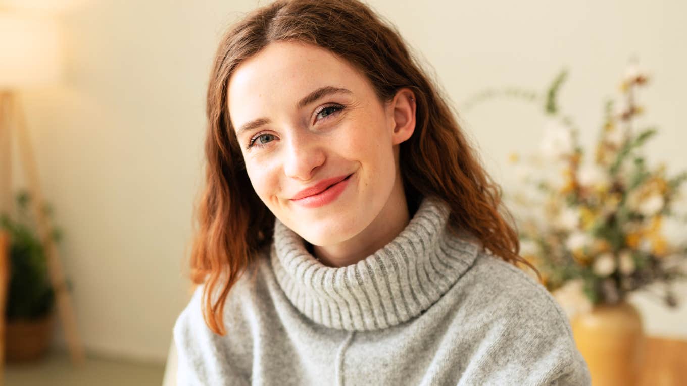 Cute young redhead woman with an engaging grin sitting perched on the end of a table looking at the camera with smiling eyes.