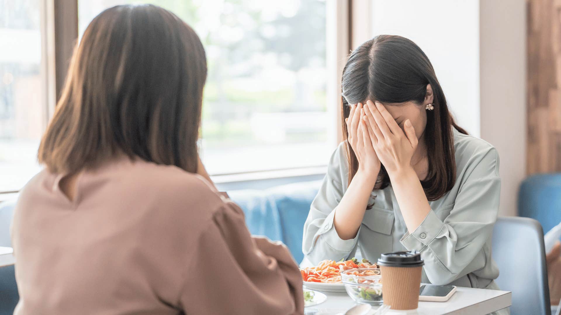 woman covering her face as she tells friend at dinner i can't believe you'd do that to me