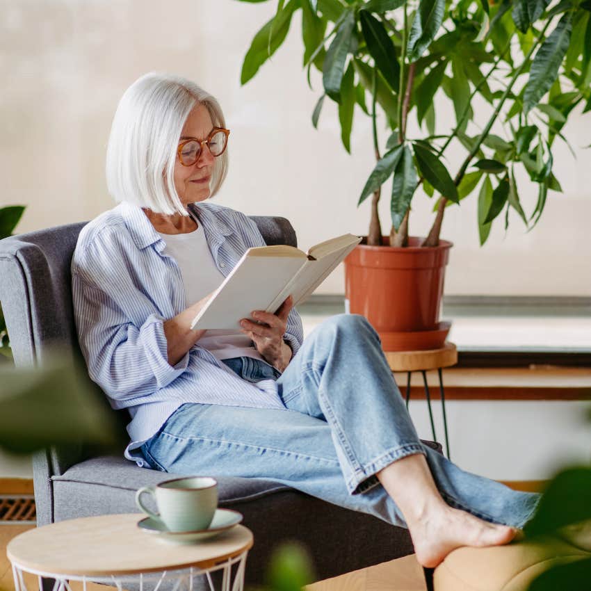 introverted woman wearing glasses reading book in her living room with her feet up