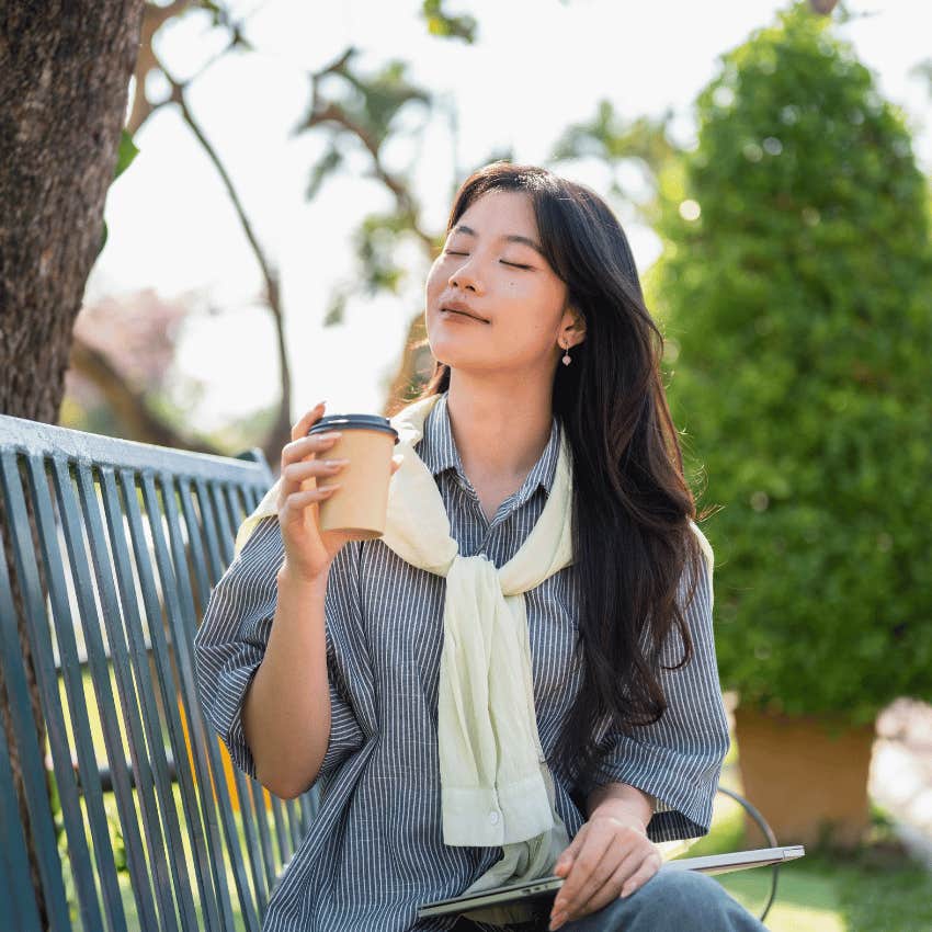 young woman sitting on park bench drinking coffee