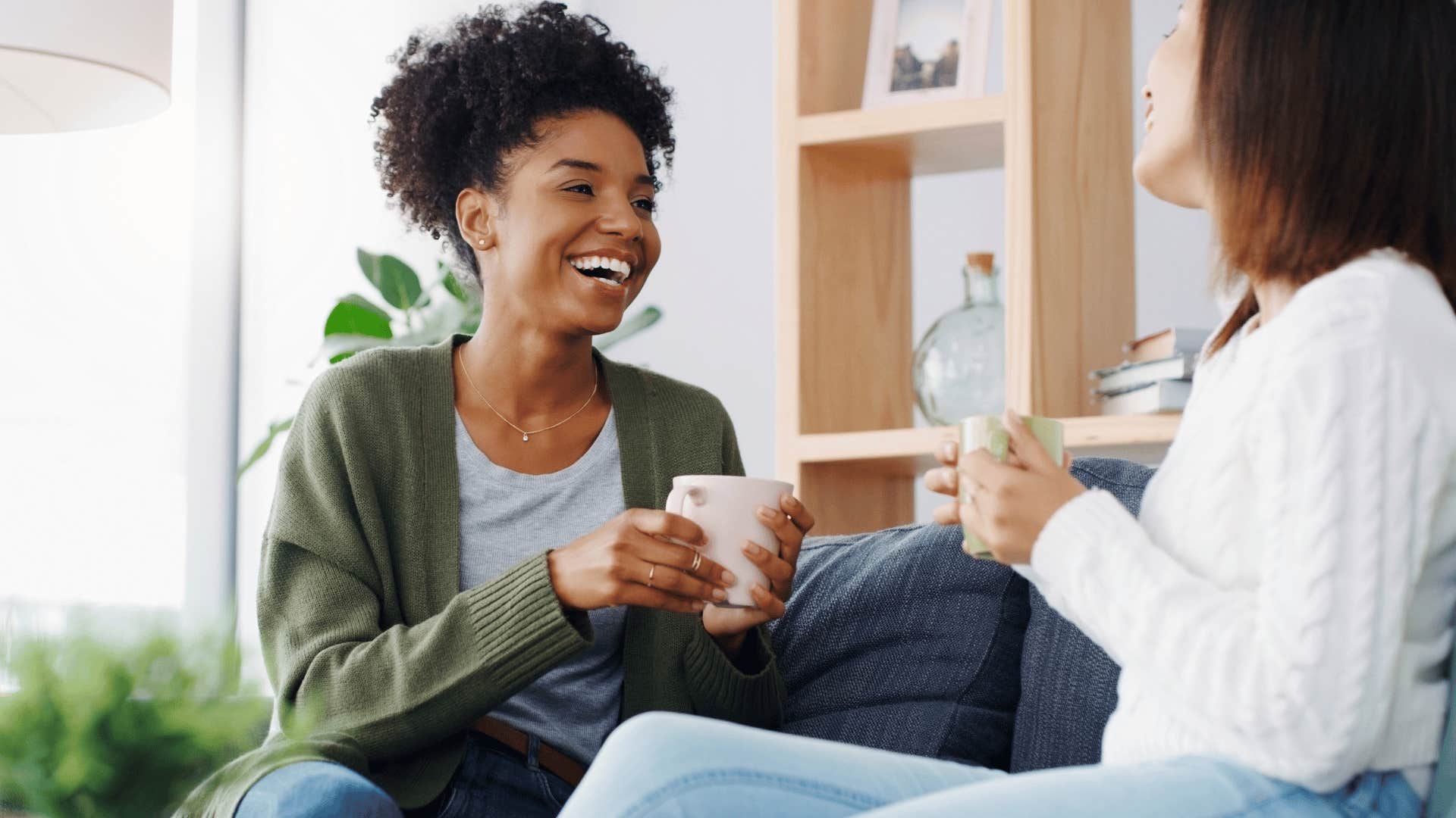 woman in green sweater saying sorry about the mess and laughing as she chats with friend on couch