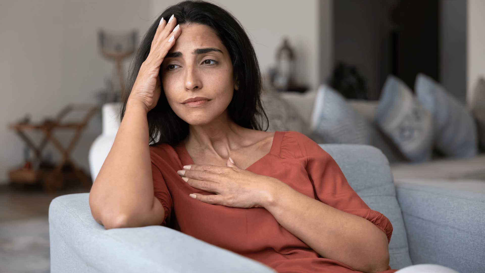 woman holding hand over heart as she thinks i can't deal with this while sitting on couch