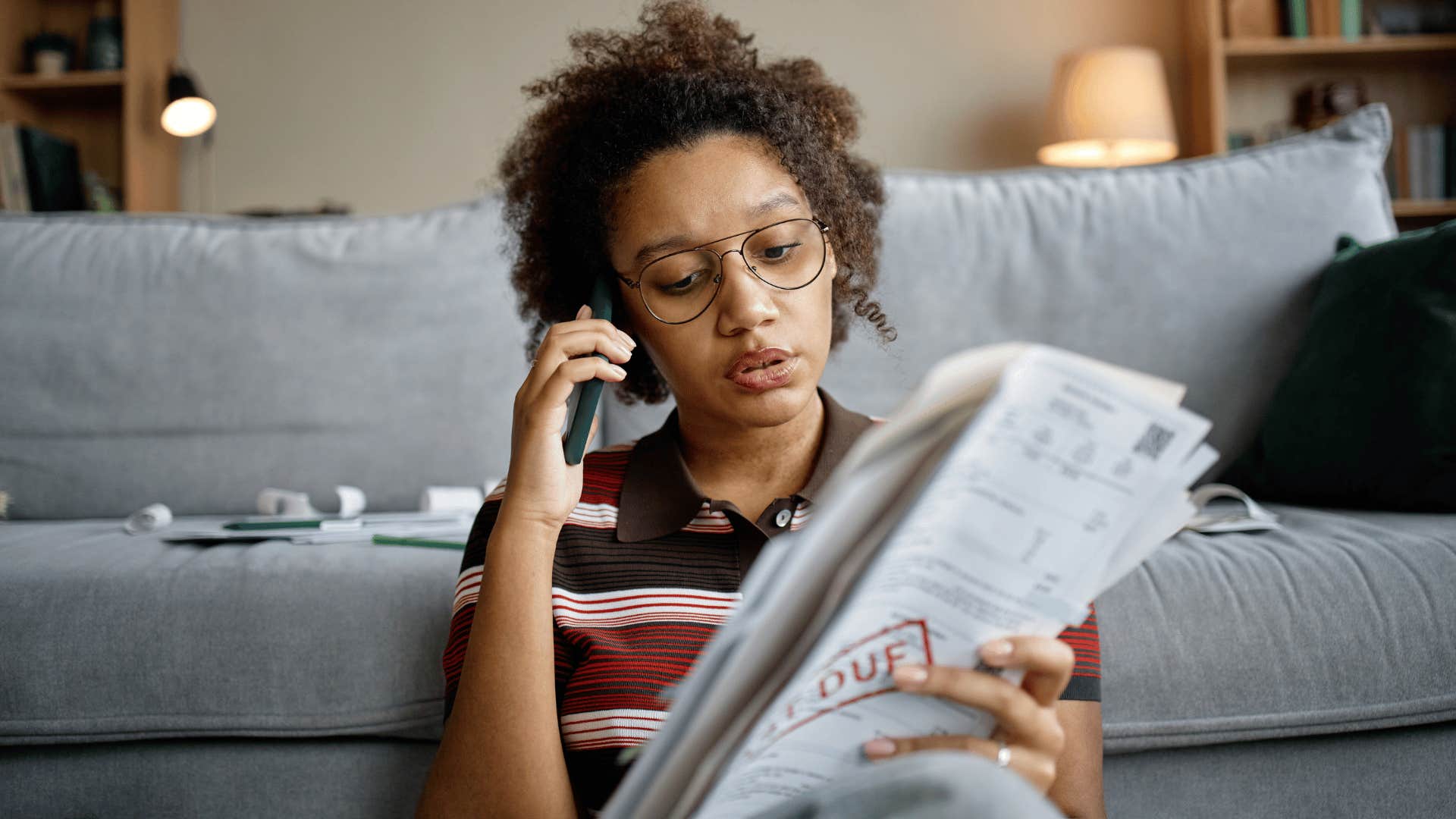 woman in stripped shirt on the phone saying i'm just trying to catch up on a few bills as she stares at documents