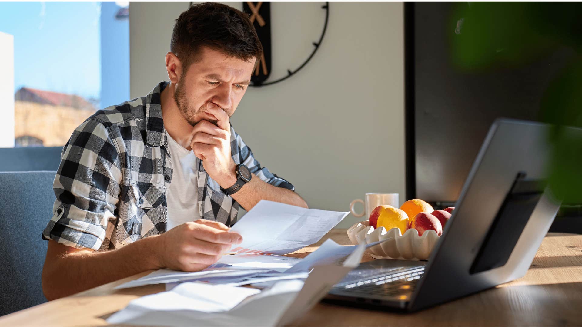 man in plaid shirt looking at bills as he thinks i'm just trying to make this paycheck stretch