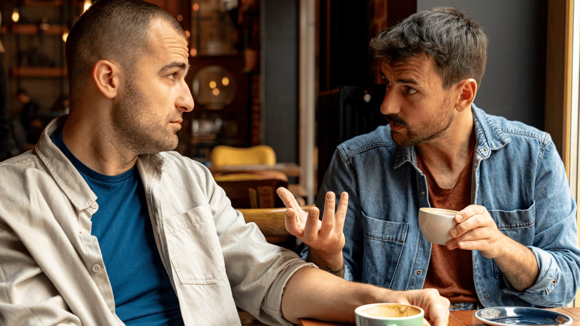 man in blue shirt annoyed at people who ask too many questions as he sits at cafe with friend
