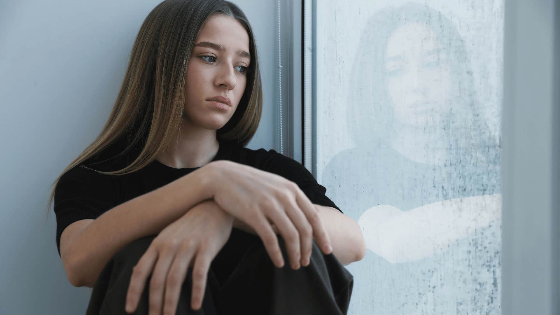 teenage girl in black shirt looking out window as she thinks life isn't fair