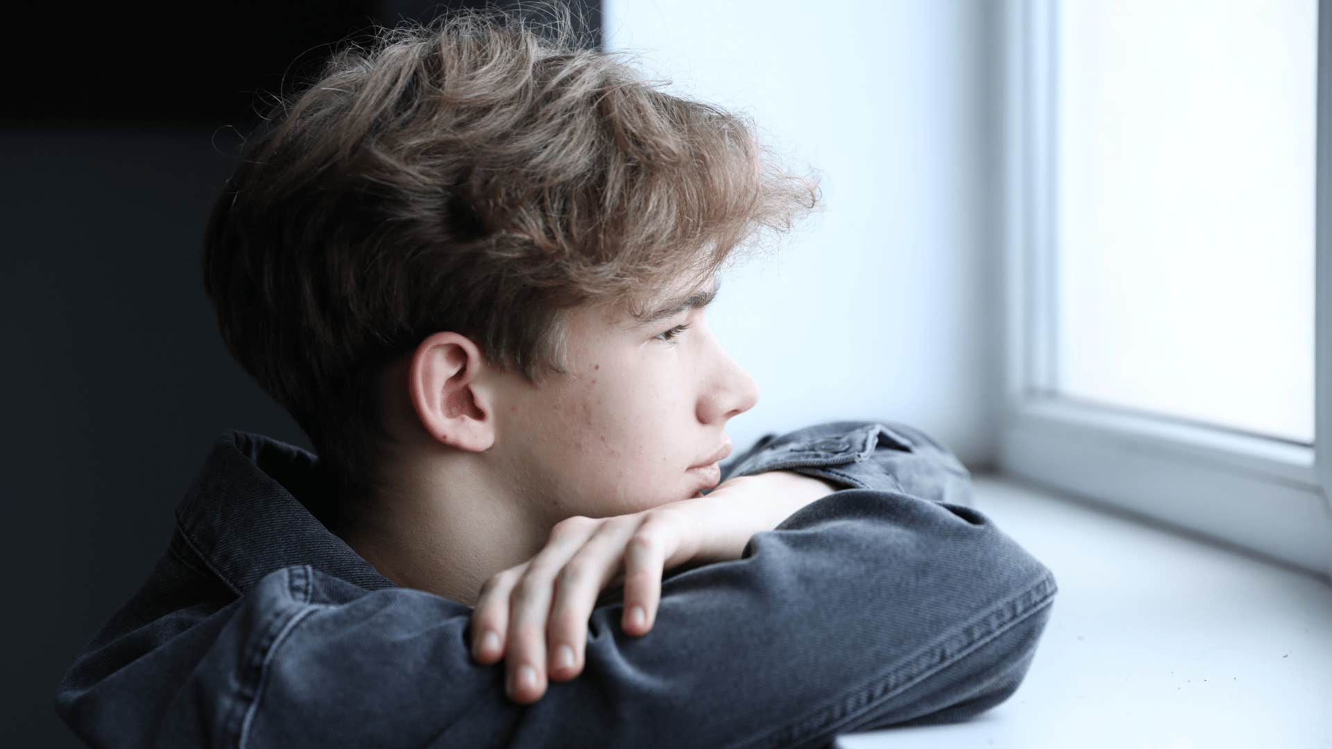 teenage boy in black shirt looking out the window upset as he's compared to siblings who could do no wrong