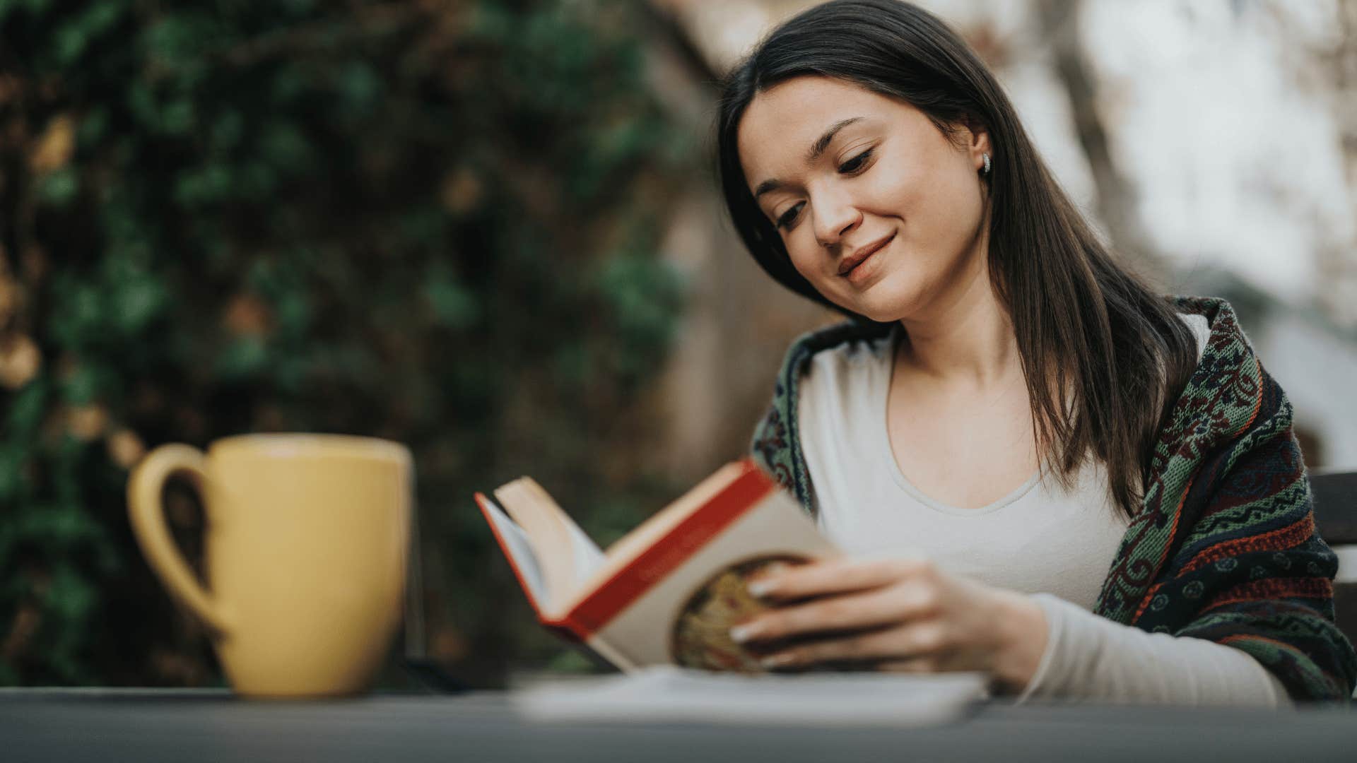 woman reading book outside with cup of coffee in front of her