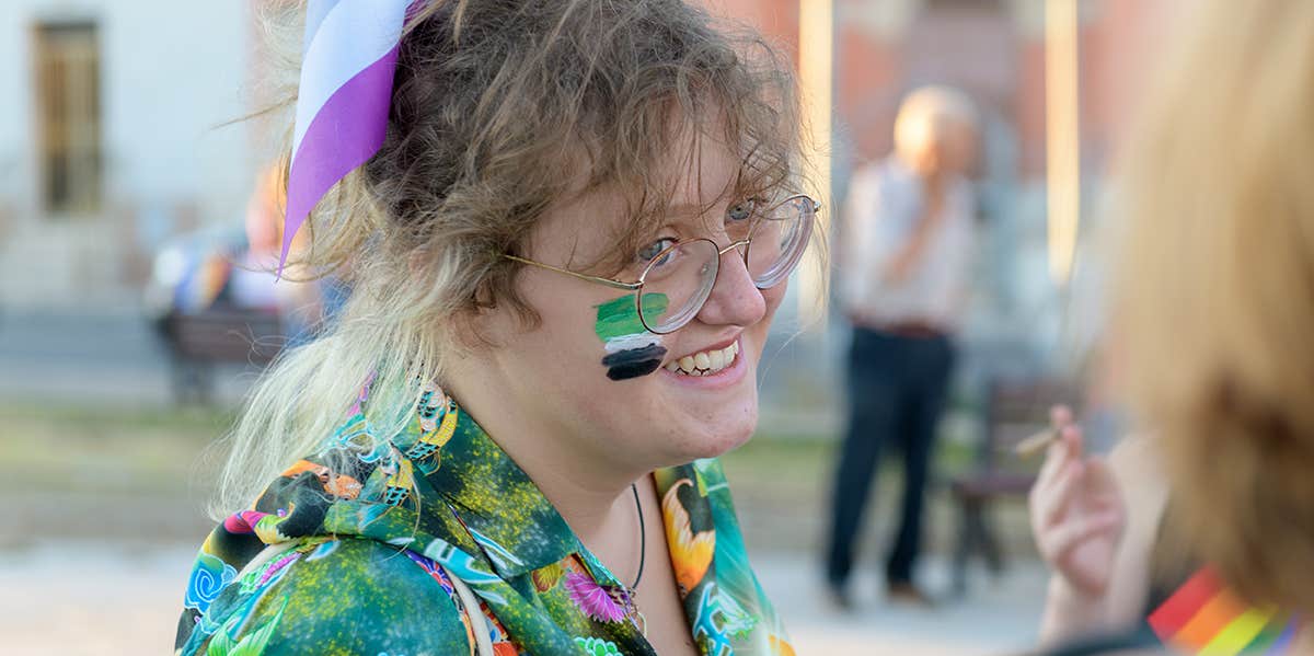 A photograph of a white person at pride, wearing a green patterned shirt, with a painting of the aromantic flag on their face.