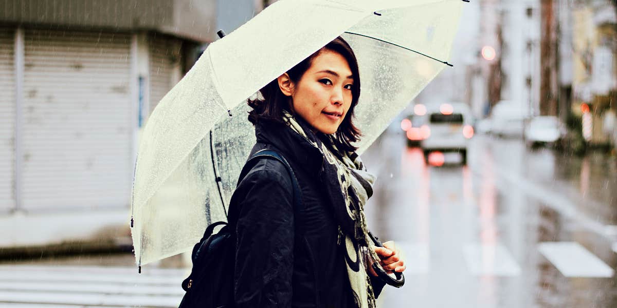 Asian woman crossing rainy street with umbrella, looking sweetly at camera