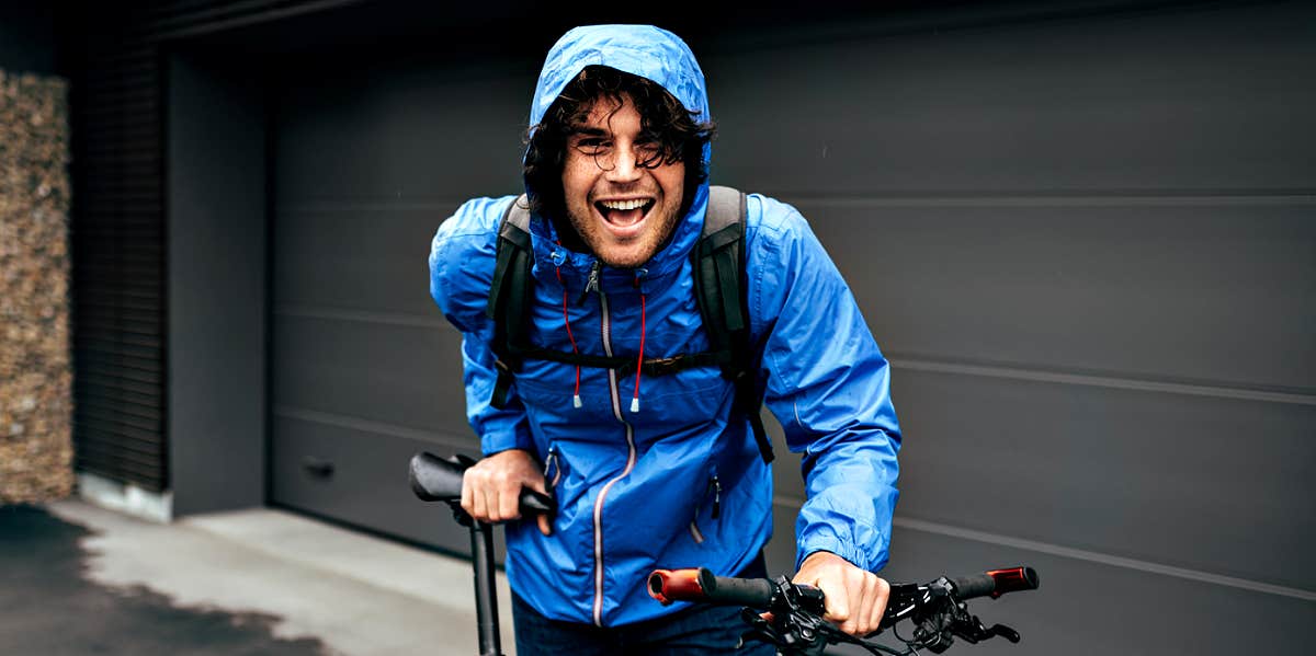 white guy with curly hair laughs into camera, wearing blue rain coat holding a mountain bike