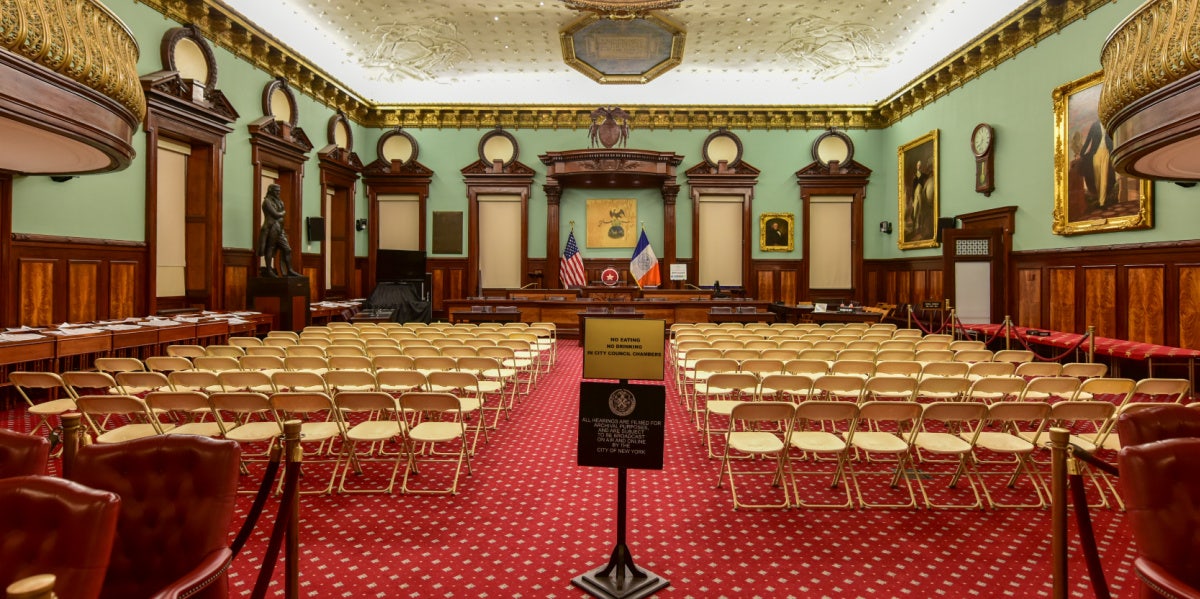 empty room where the NYC city council usually meets, filled with empty chairs and empty dais
