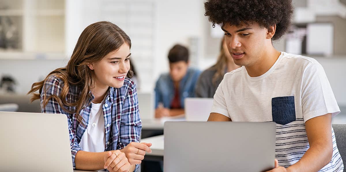 A photograph of a two students in a classroom, with the male student showing his laptop to a female student.