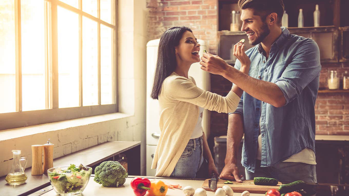 Beautiful young couple is feeding each other and smiling while cooking in kitchen at home