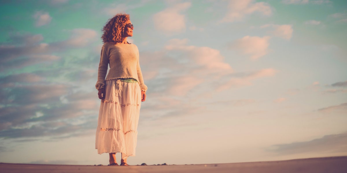 15 Ways Women's Bodies Change As We Age beautiful aging woman with curly brown hair stands on sand in front of vibrant sky