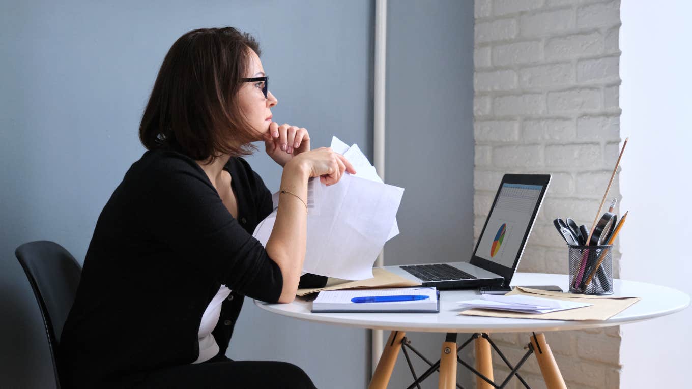 sad middle-aged woman looking out window with papers and laptop computer sitting at home at the table.