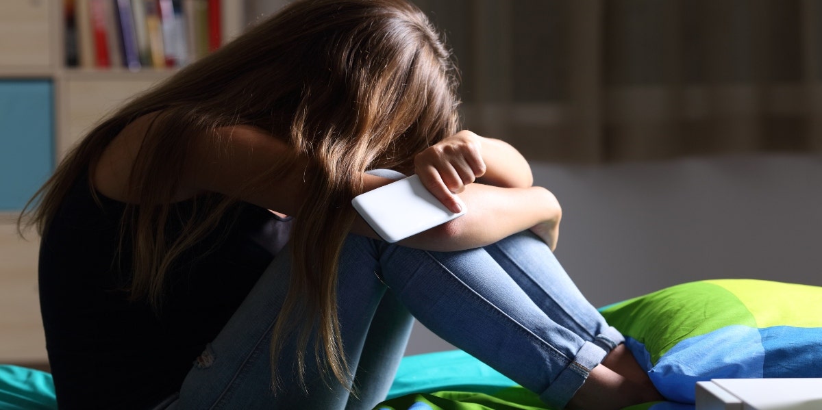 The April 24 'National Rape Day' Hoax On TikTok Explained scared woman hugging her knees while sitting on a bed with her phone