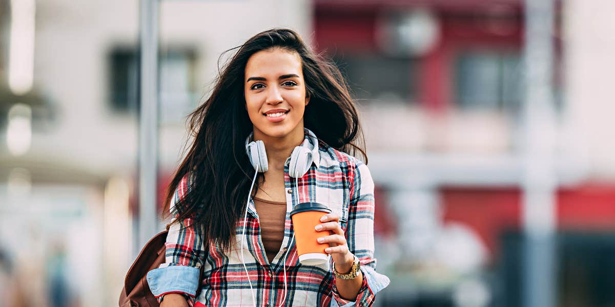 Young Latina woman crossing a city street with coffee and headphones