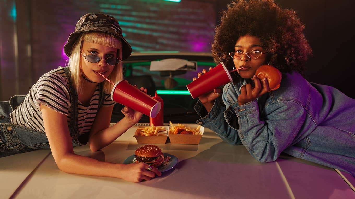 Two women eating takeout food on the hood of the car in the parking lot.