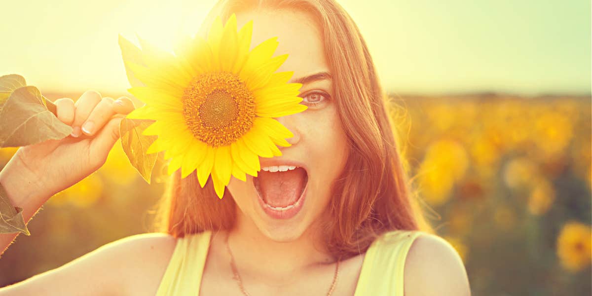 Young woman in a sunflower field smiling behind a flower, sunlit from behind
