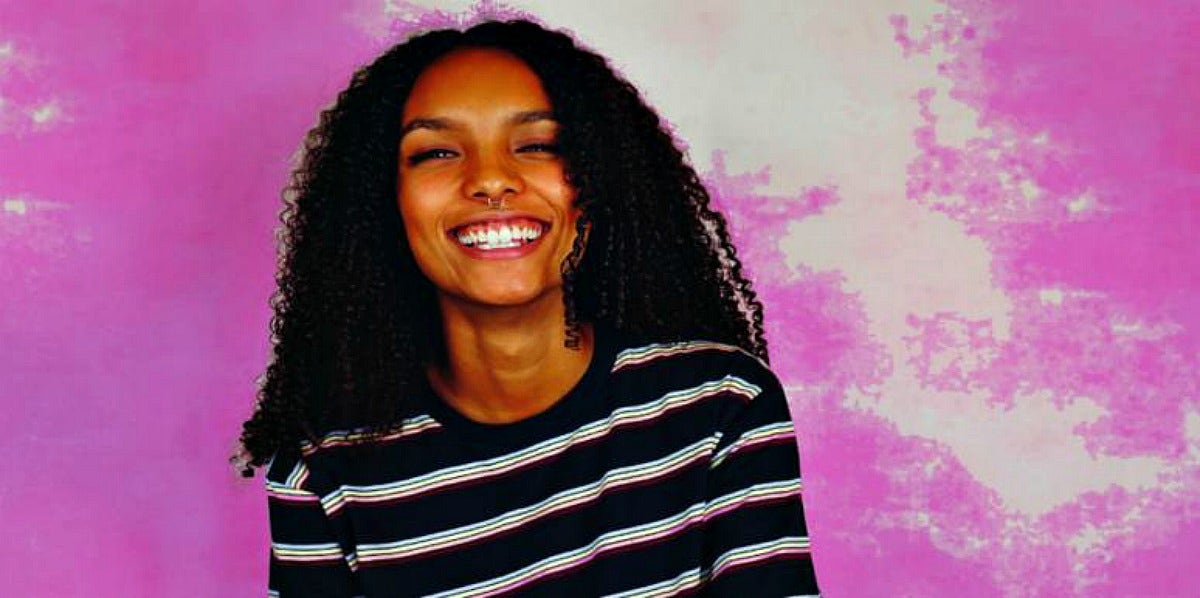 twenty-something woman with long, curly natural hair smiles in front of a purple background