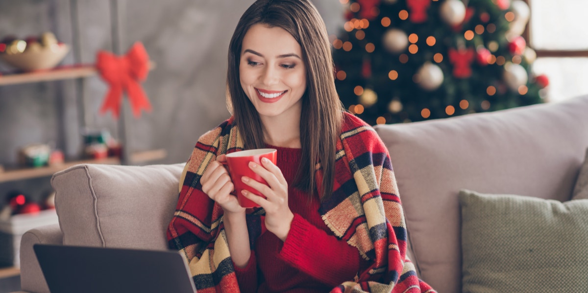 20 Best Online Holiday Cards Of 2021 — Christmas, Hanukkah, New Year's & More woman drinking hot chocolate looking at laptop screen