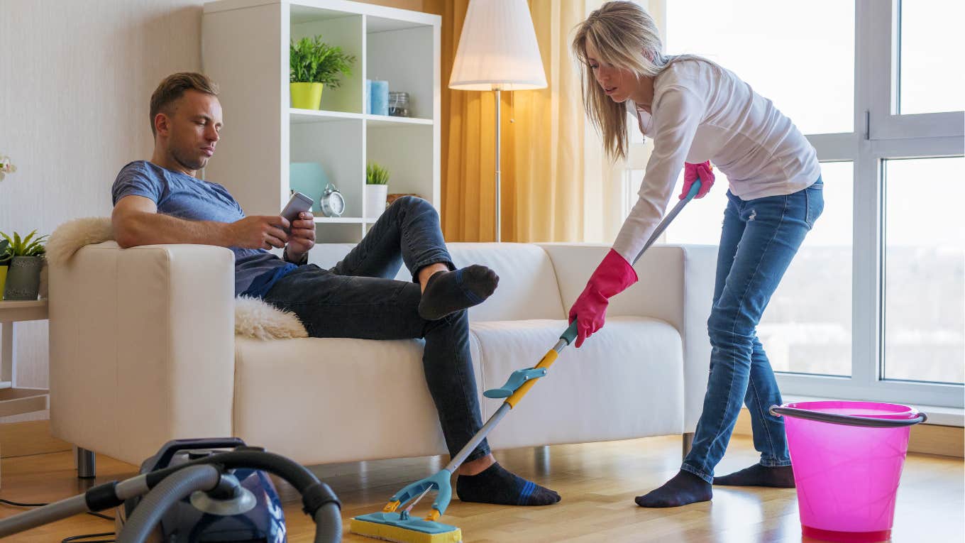 Woman cleans the house, while her boyfriend sits on the couch. 