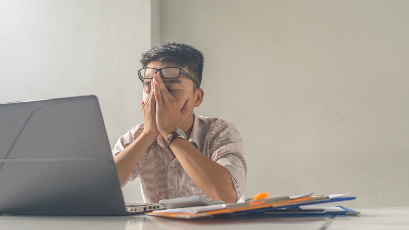 Burnout Coach Advises Salaried Employees To Go Home After 40 Hours tired employee rubbing face while working in front of a laptop at a desk