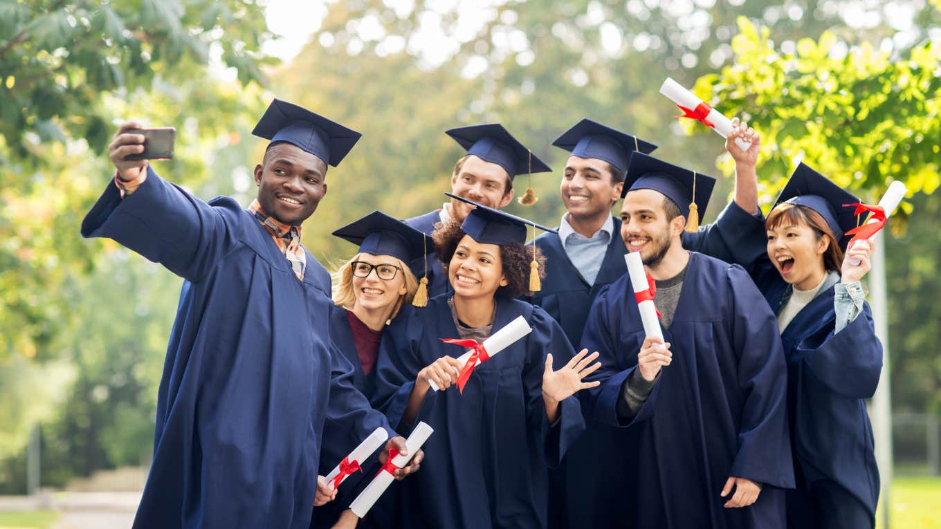 group of happy students in mortar boards and bachelor gowns with diplomas taking selfie outdoors