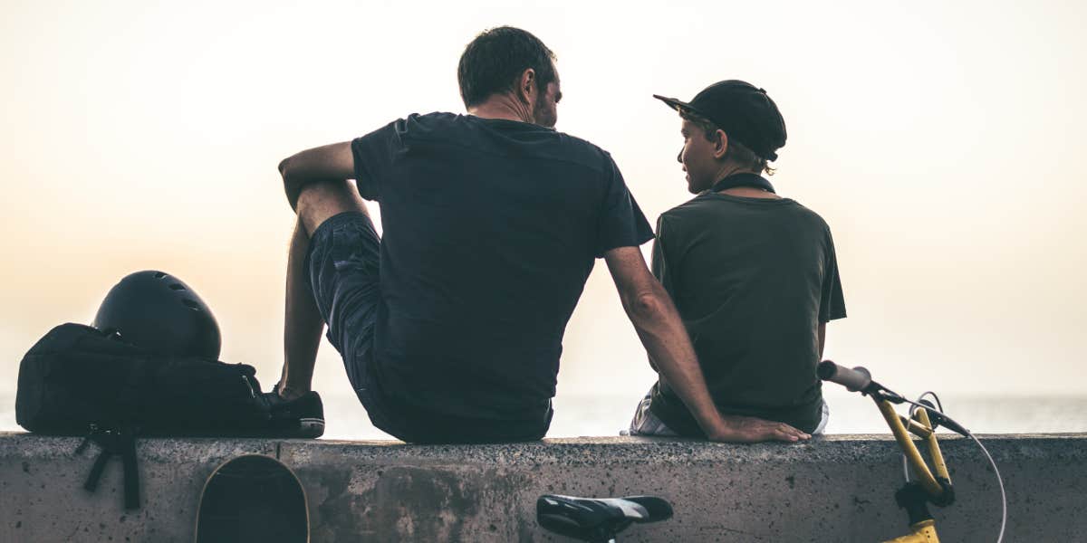 Father and son, backlit by sunset, sit on a wall with a bike and skateboard 