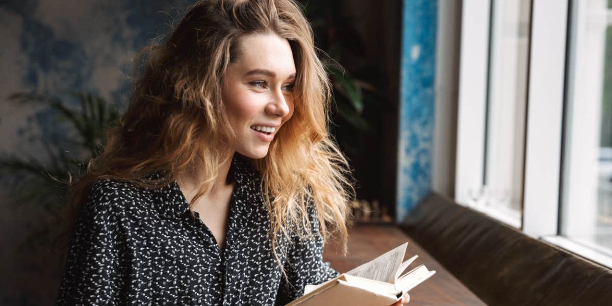 young woman with long blonde hair reads a book by a window, coffee cup nearby