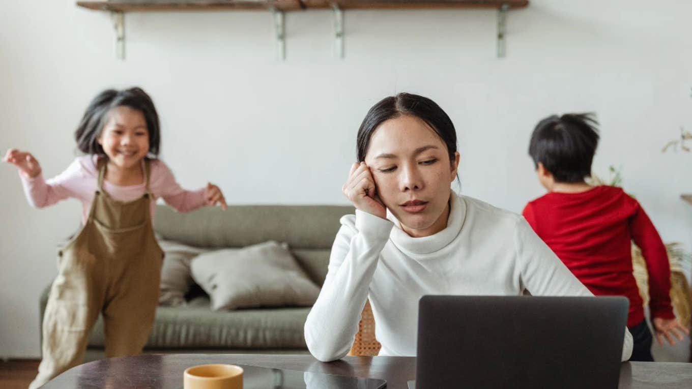 stressed woman using laptop while two young kids play in the background