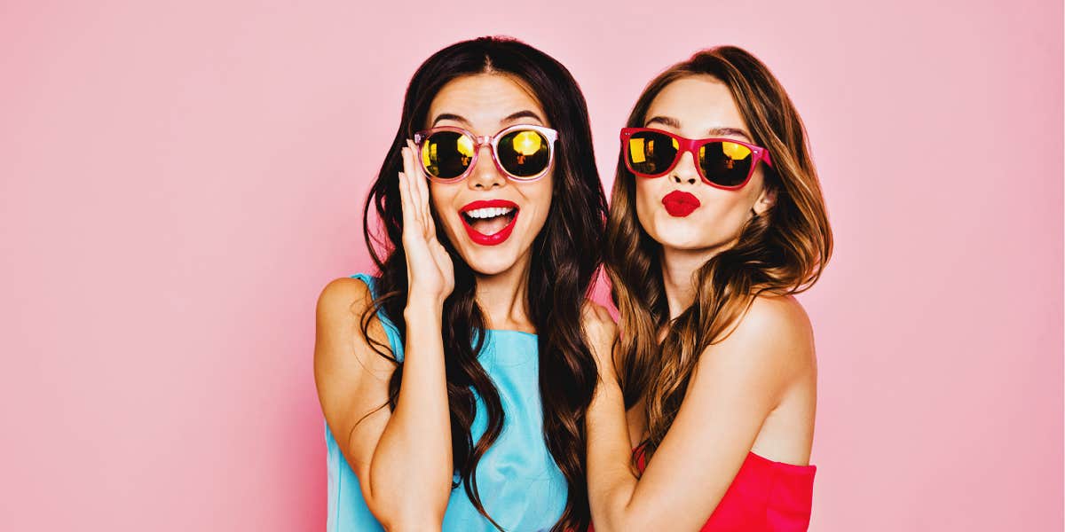 two young women in bright dresses and sunglasses, pink background