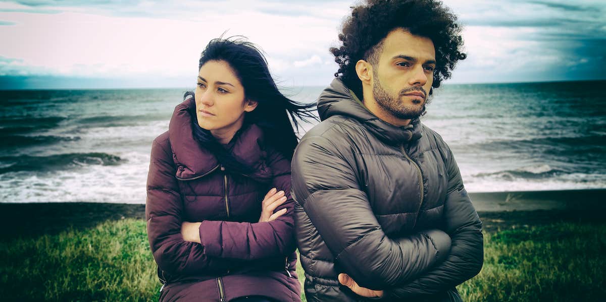 Upset couple stand with ocean behind them. Their arms are crossed, anger paints their expressions