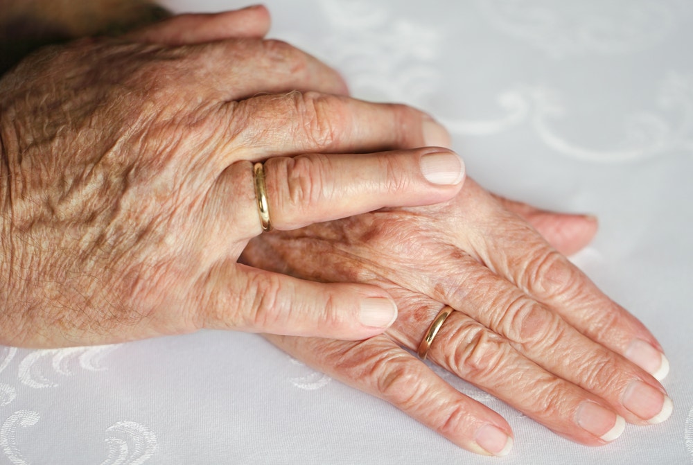 An elderly couple holds hands with wedding bands on their fingers.