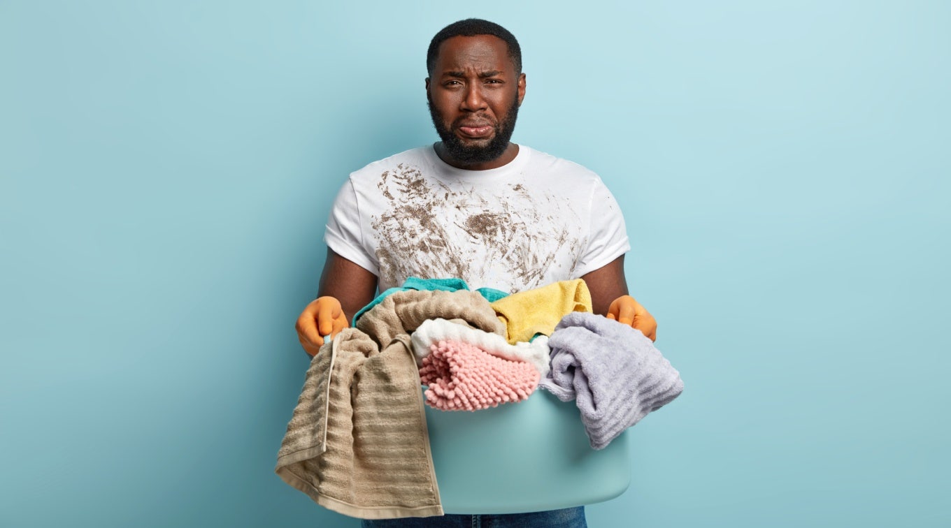 Sad pity African American husband overloaded with housework, wears dirty clothes and gloves has duties to wash linen, cries from depression and puzzlement, stands over blue background. Too much work Man crying while holding laundry hamper.